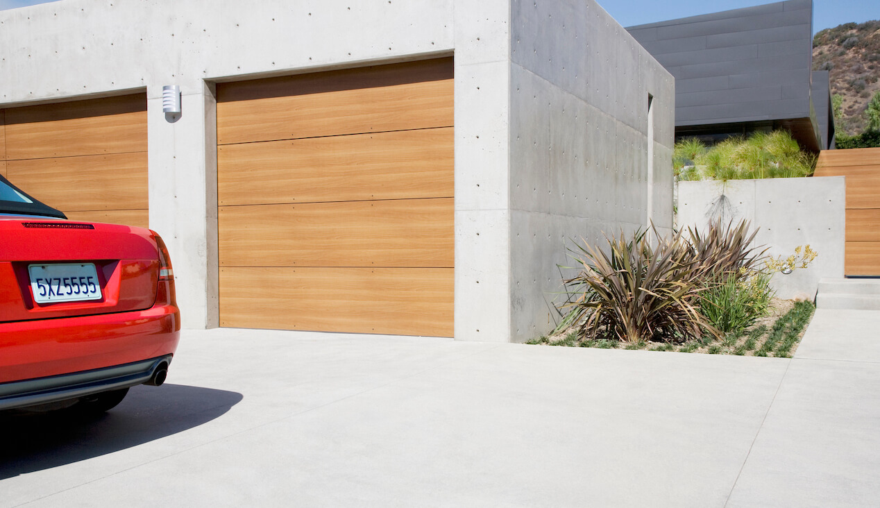 concrete driveway and garage with wooden garage door and partial view of red car parked nearby